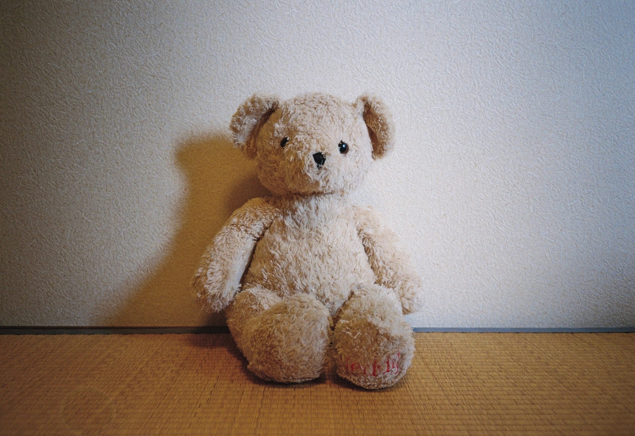 A worn cream teddy bear with a red-stitched paw sits against a textured white wall on a tatami floor.