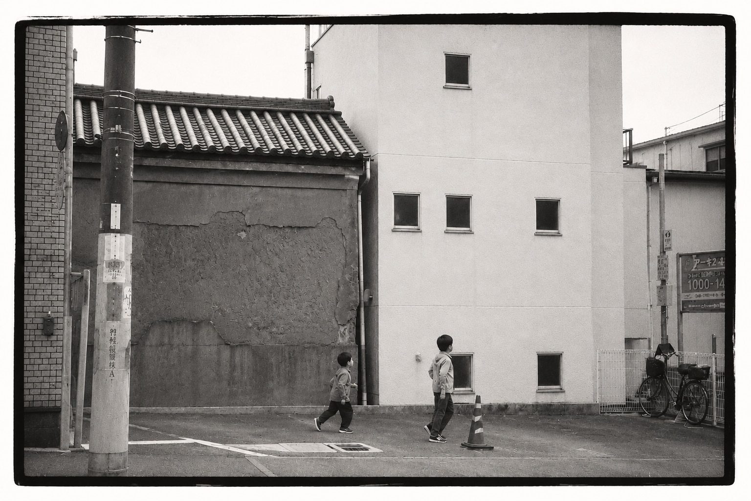 Two young children walking away across a paved lot, past a traffic cone, in front of a plastered building with small square windows and an old tiled-roof wall; a bicycle leans against a fence.