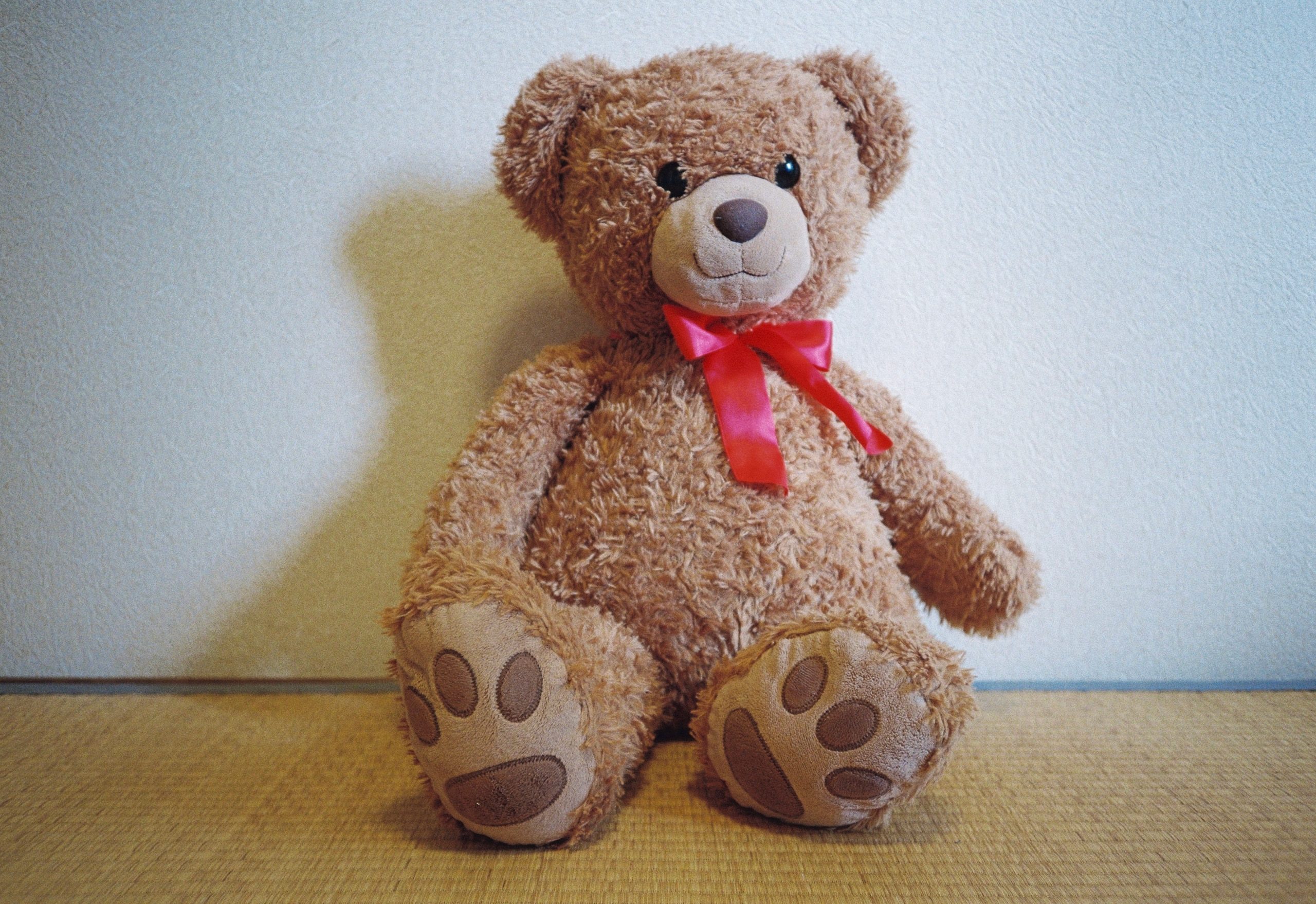 A brown teddy bear with a red satin bow and stitched paw pads sits upright against the same white wall.