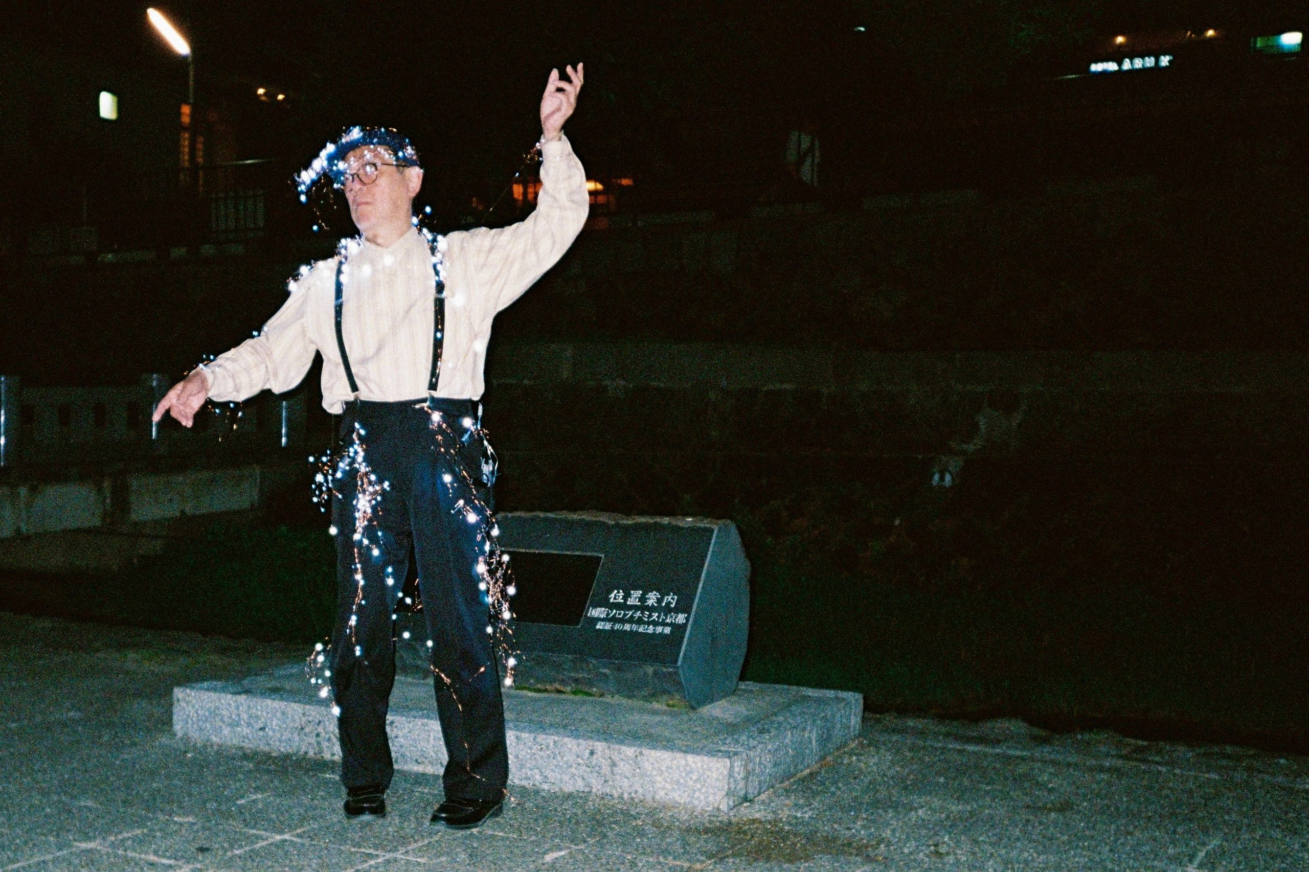 A man dances at night beside a stone monument, strung with small lit fairy lights.