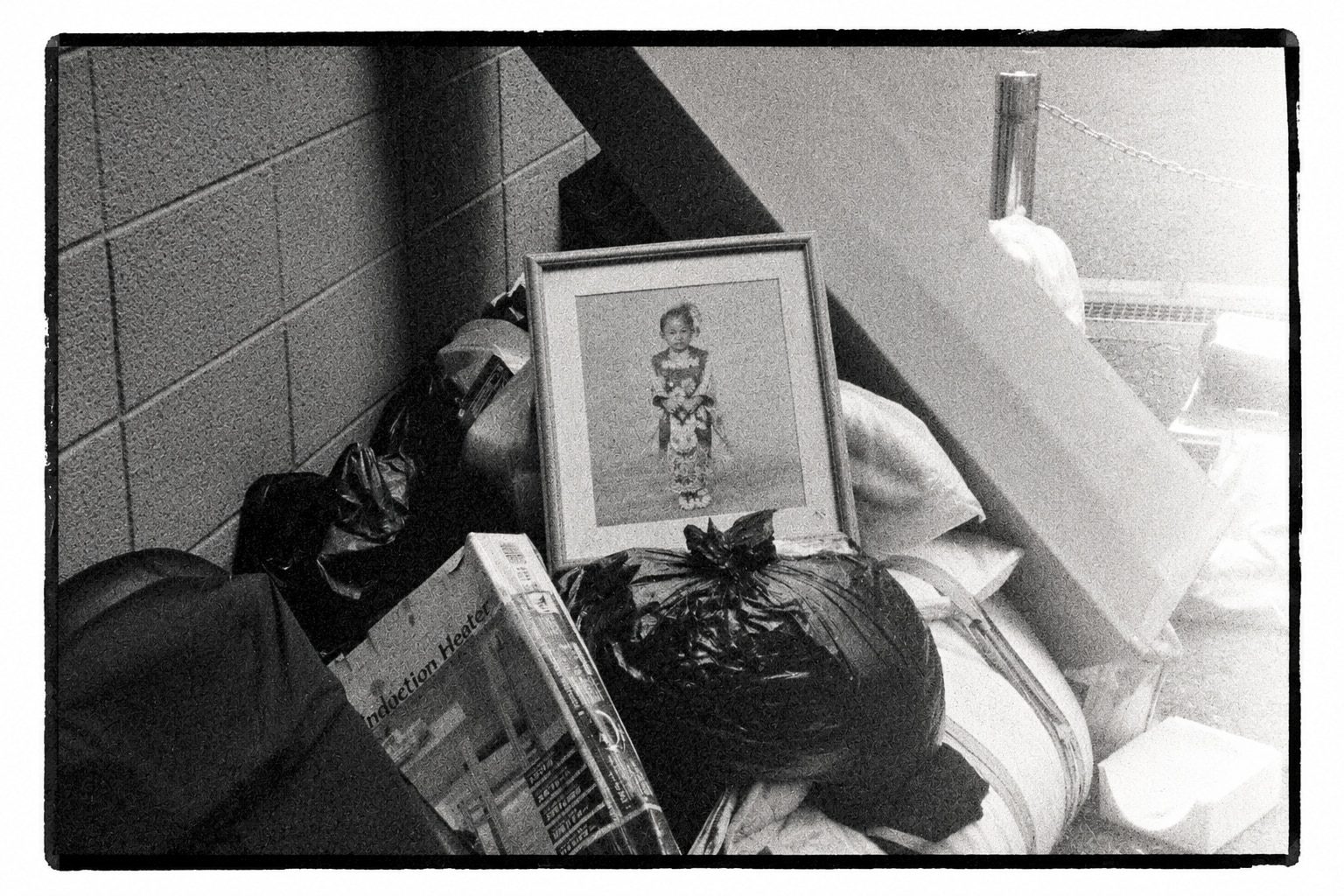 A framed portrait of a child in kimono propped on black garbage bags and bedding stacked against a tiled wall.