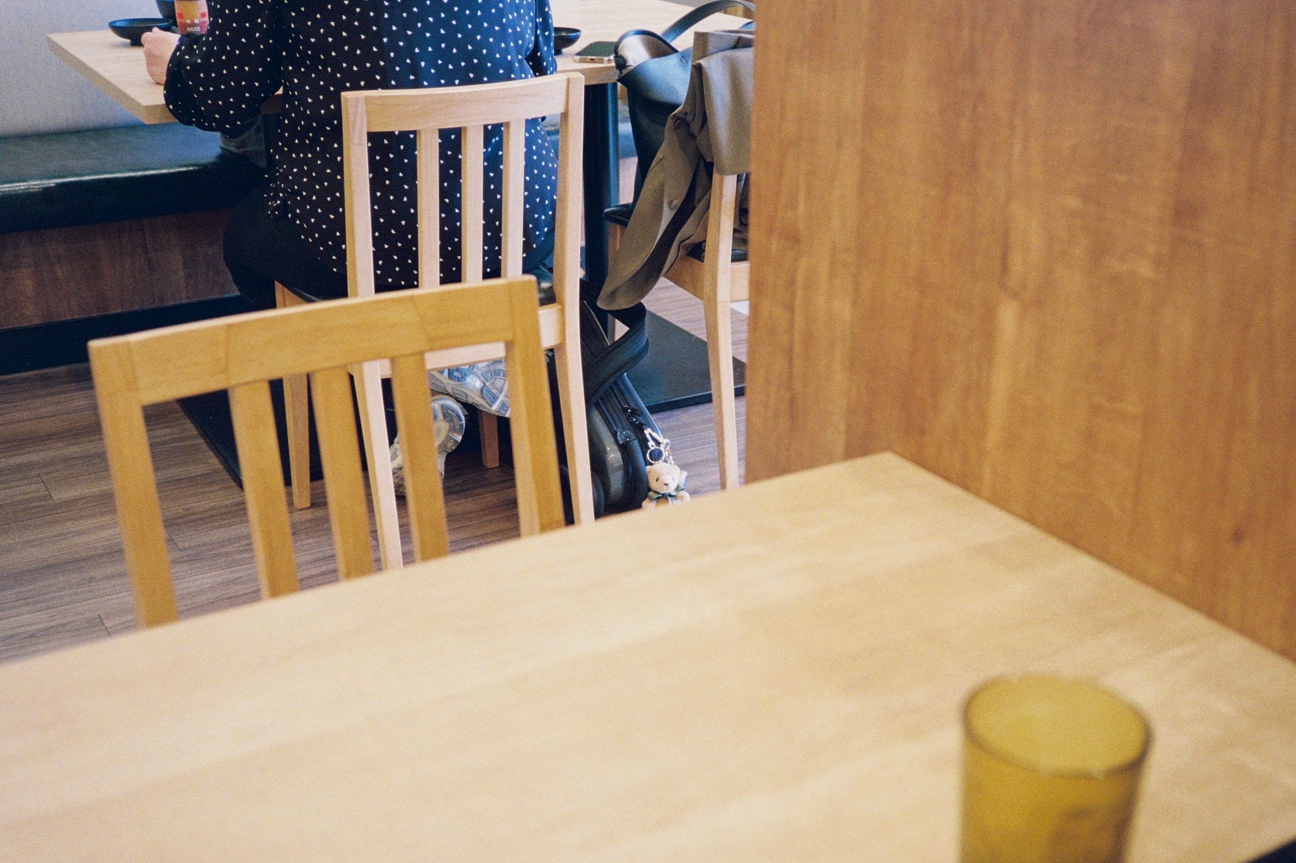 A restaurant interior with wooden tables and chairs, a person seated in the background, a glass in the foreground.