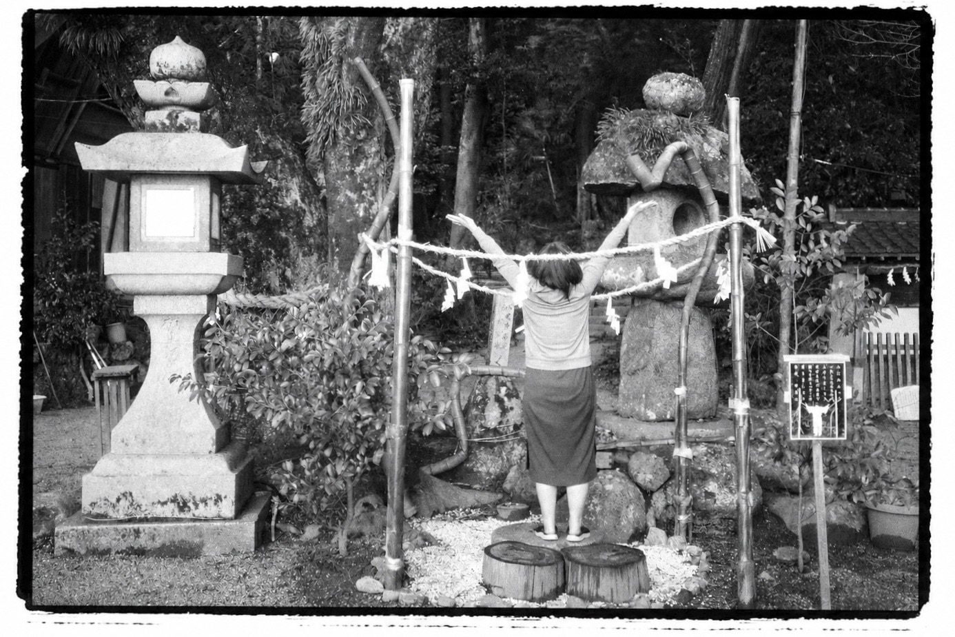 A woman seen from behind, arms raised, standing between a stone lantern and a stacked-stone shrine marker strung with a shimenawa rope.