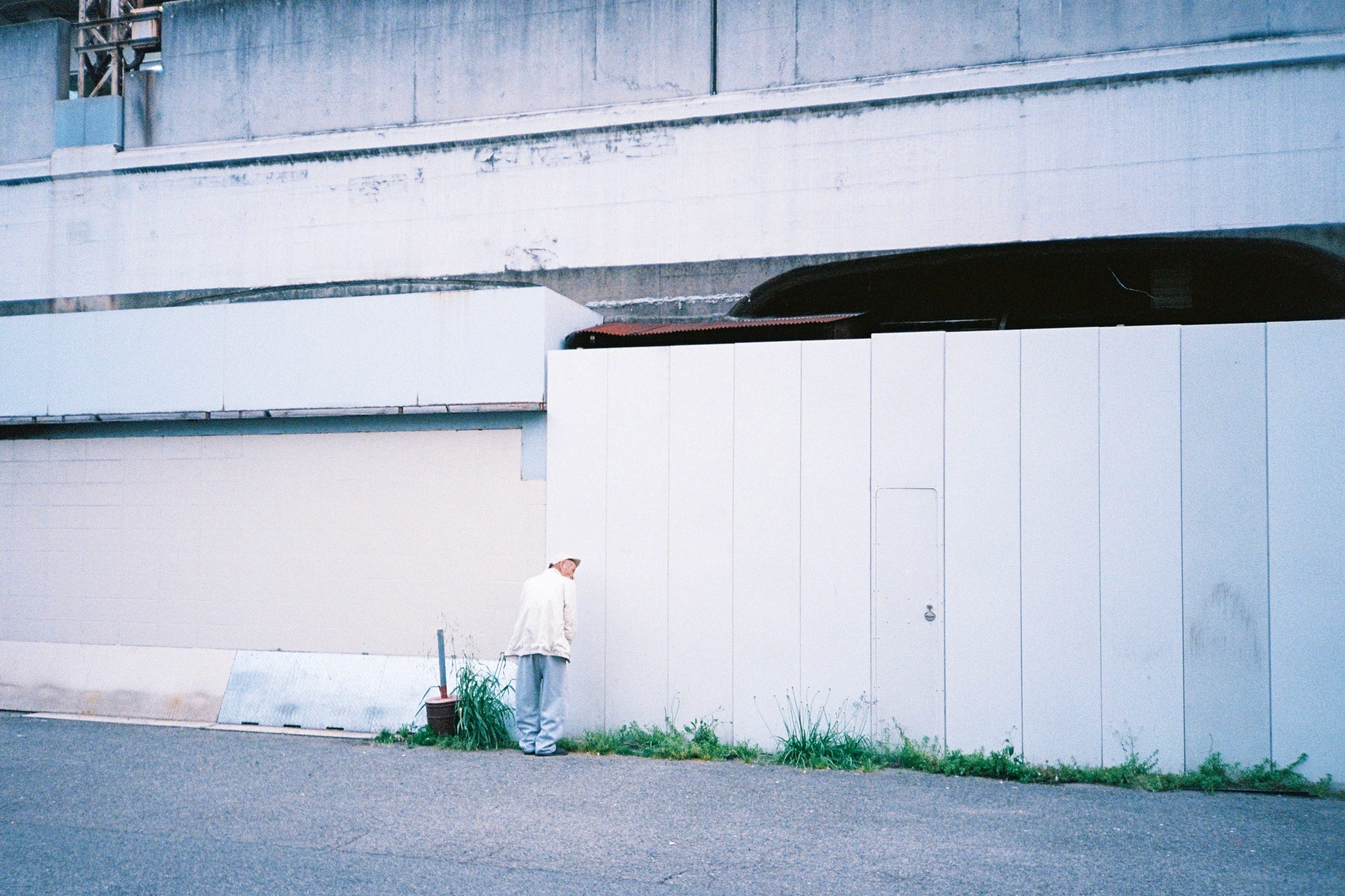 A man leans face-first against a large blank white wall beneath an overpass.