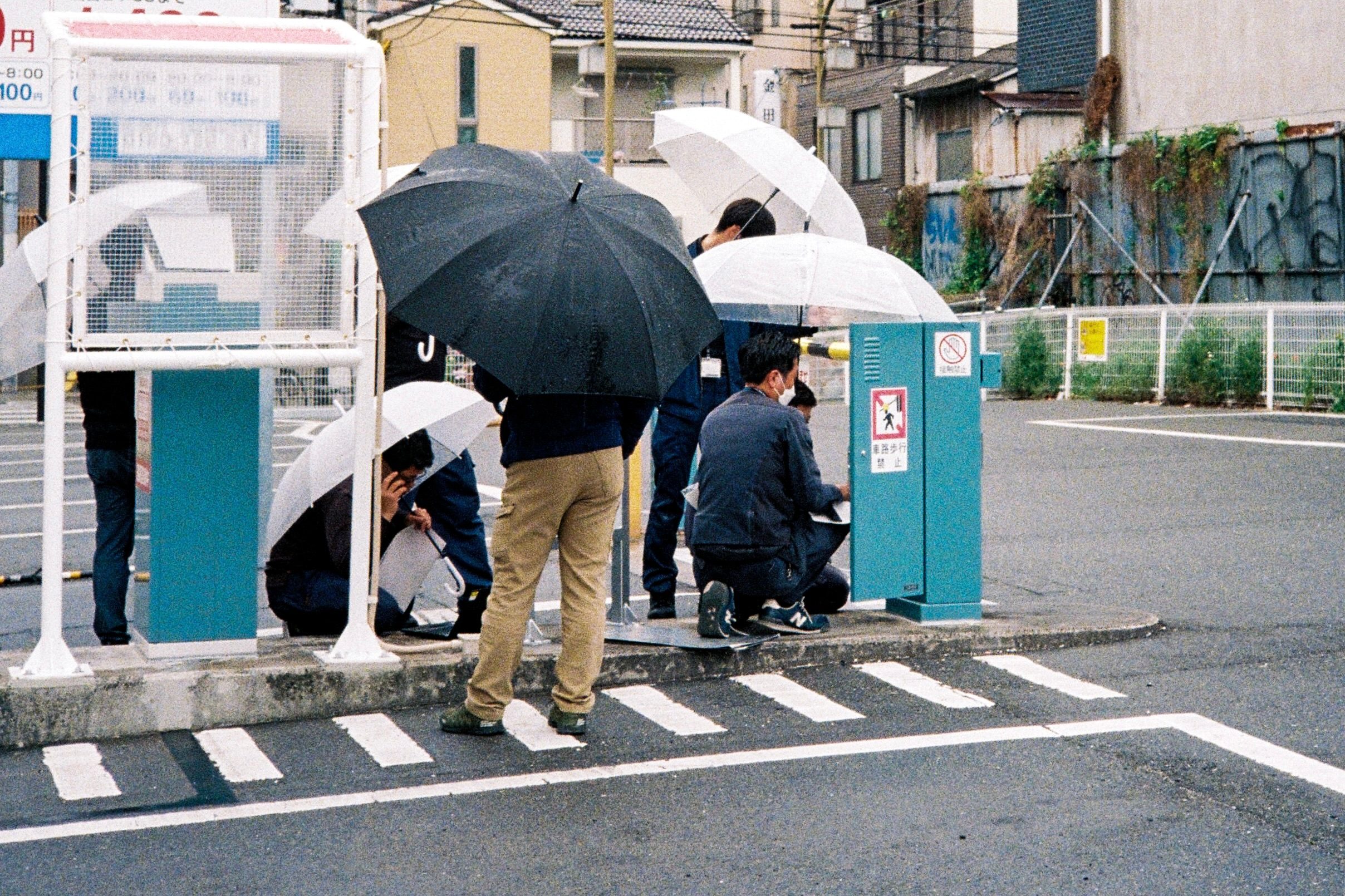 Several people crouch under umbrellas around a roadside utility box in the rain.