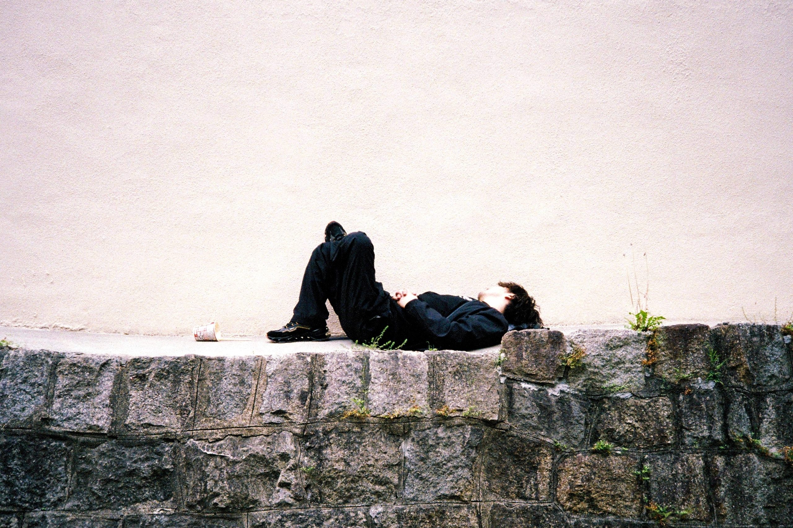 A figure lies still on a low stone wall against a vast blank stucco facade.