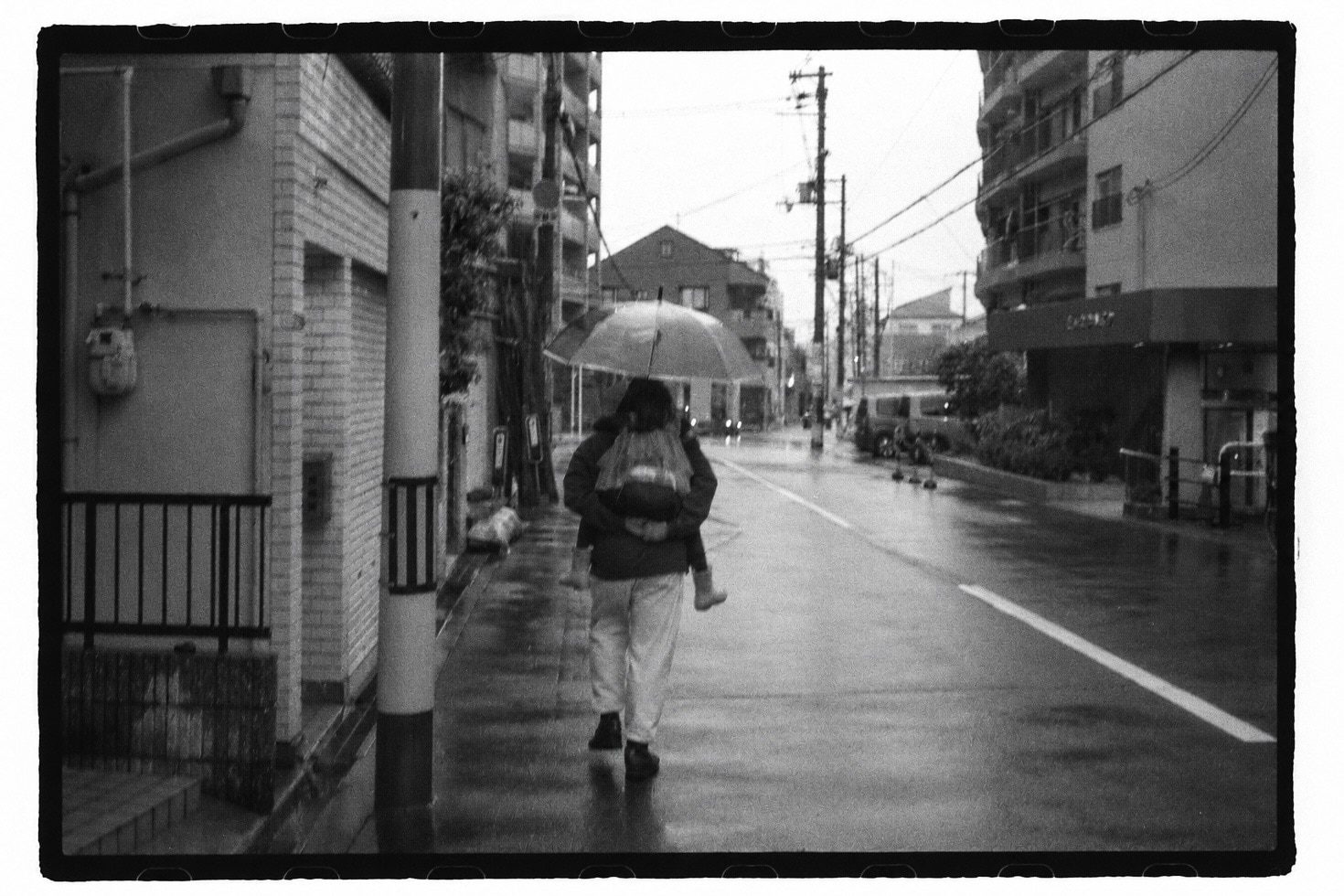 A person under an umbrella carrying a child on their back, alone on a wet street between apartment buildings.