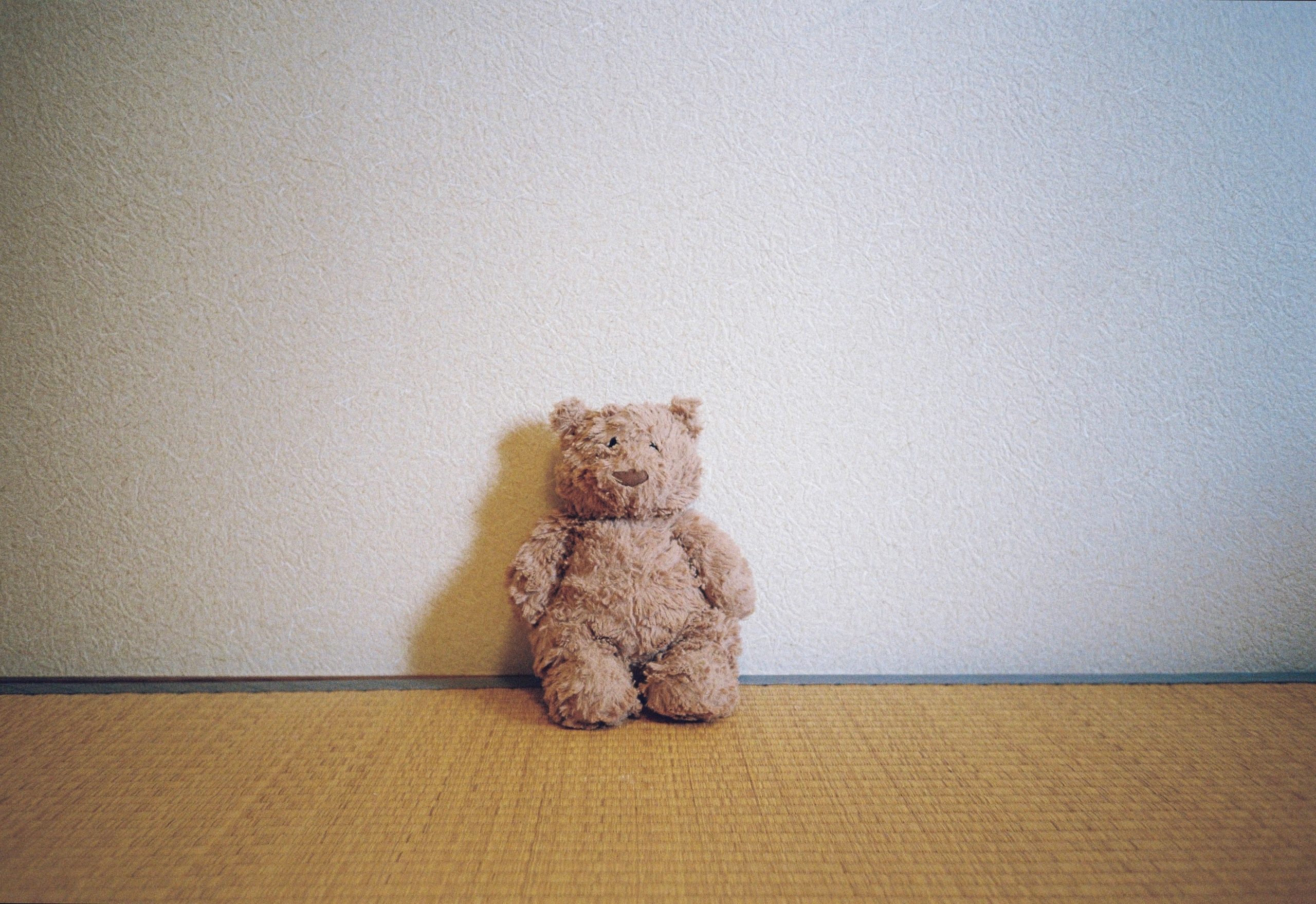 A small tan bear with a torn ear and a stitched smile sits alone in the corner of the room.