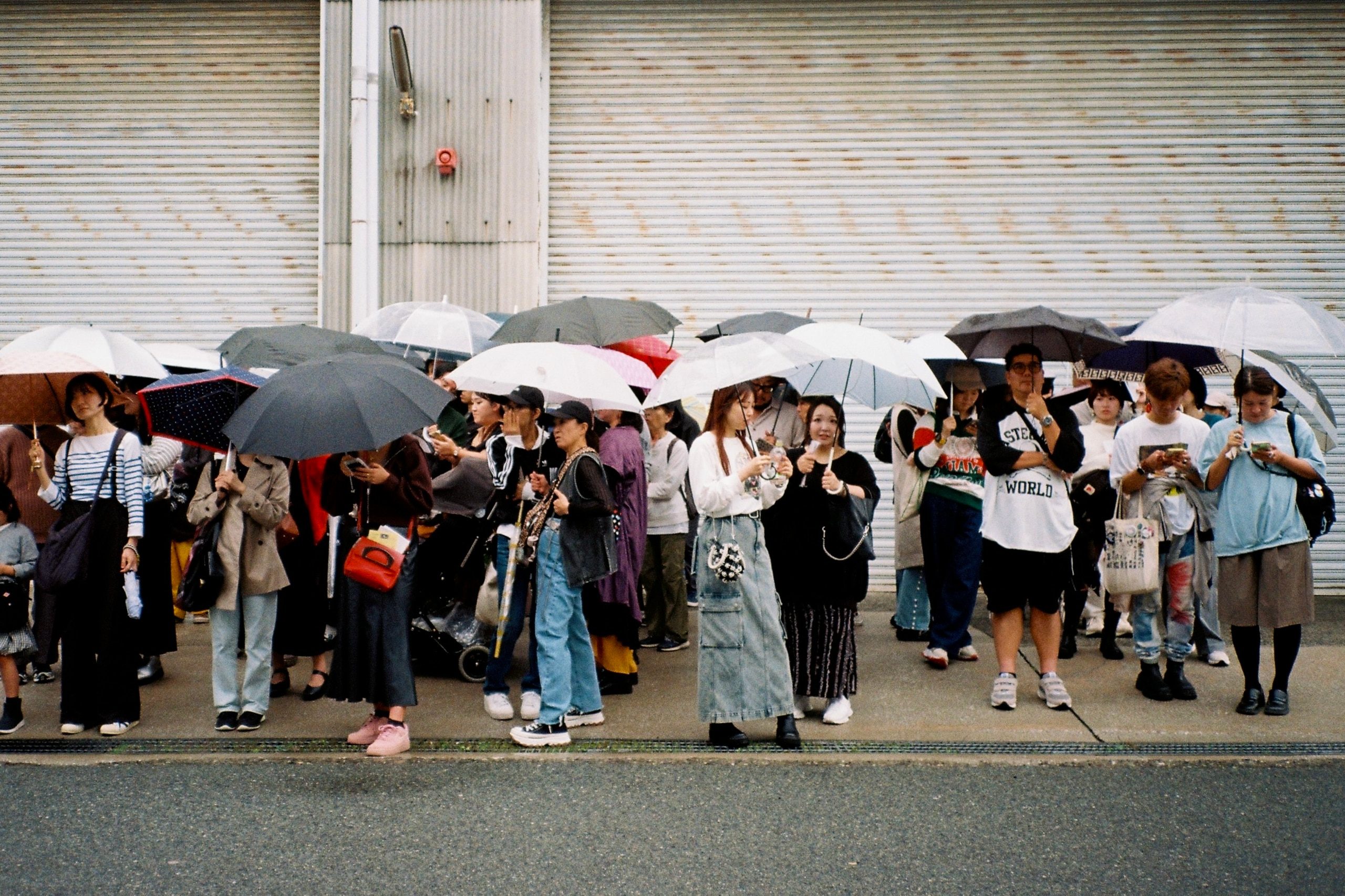 A crowd of people holding umbrellas stands together in front of a shuttered building.