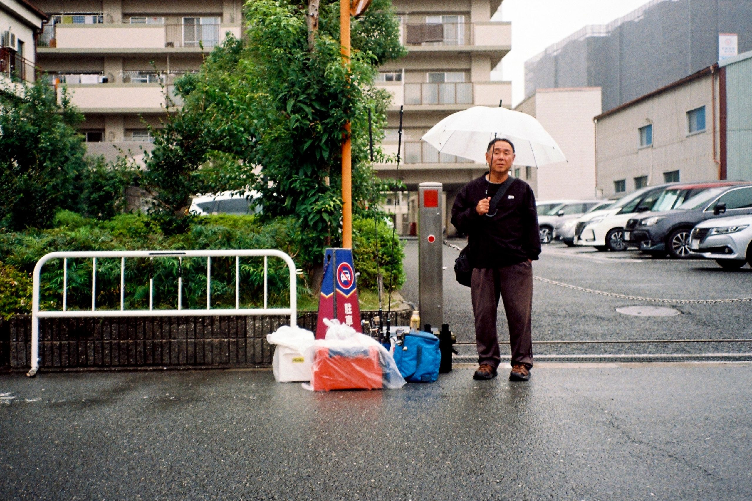 A man stands holding an umbrella in a wet parking lot beside fishing gear and bags on the ground.