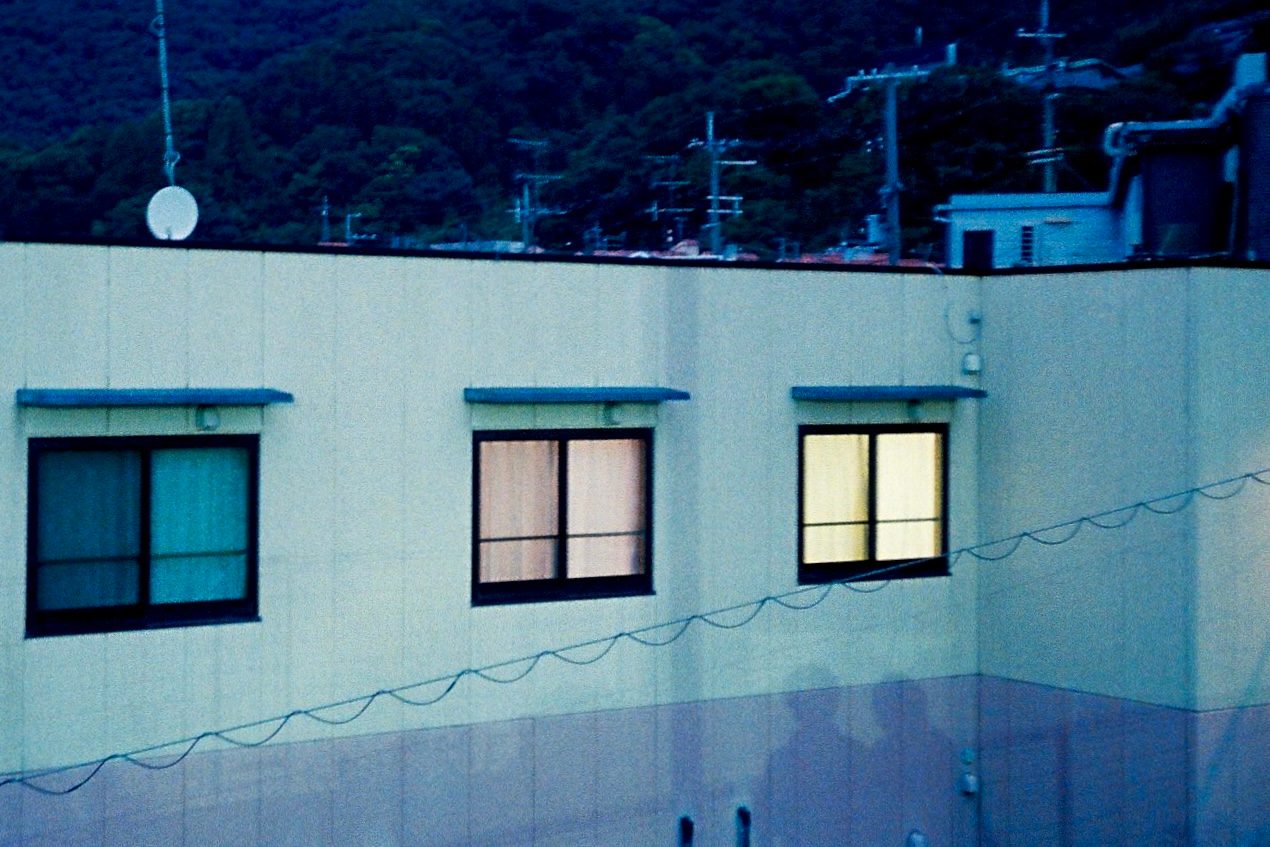 A building facade at dusk with three windows; two glow warm from within, one stays dark, hills behind.