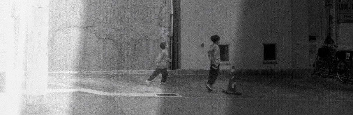 Panoramic shot of two children running across sunlit pavement cut by a band of shadow, bicycles parked beside a building.