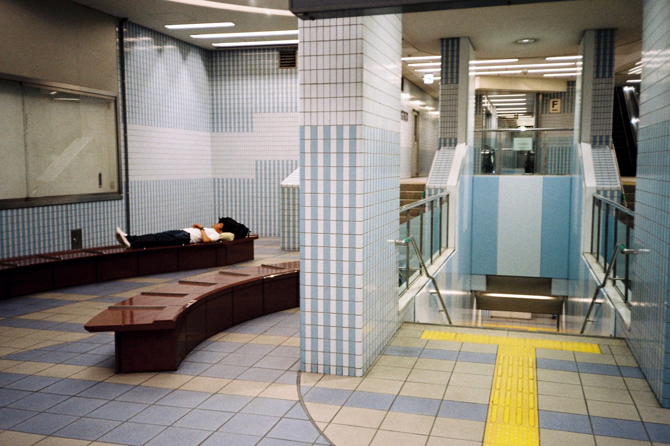 A person asleep on a bench in a hollow, tiled underground station corridor.