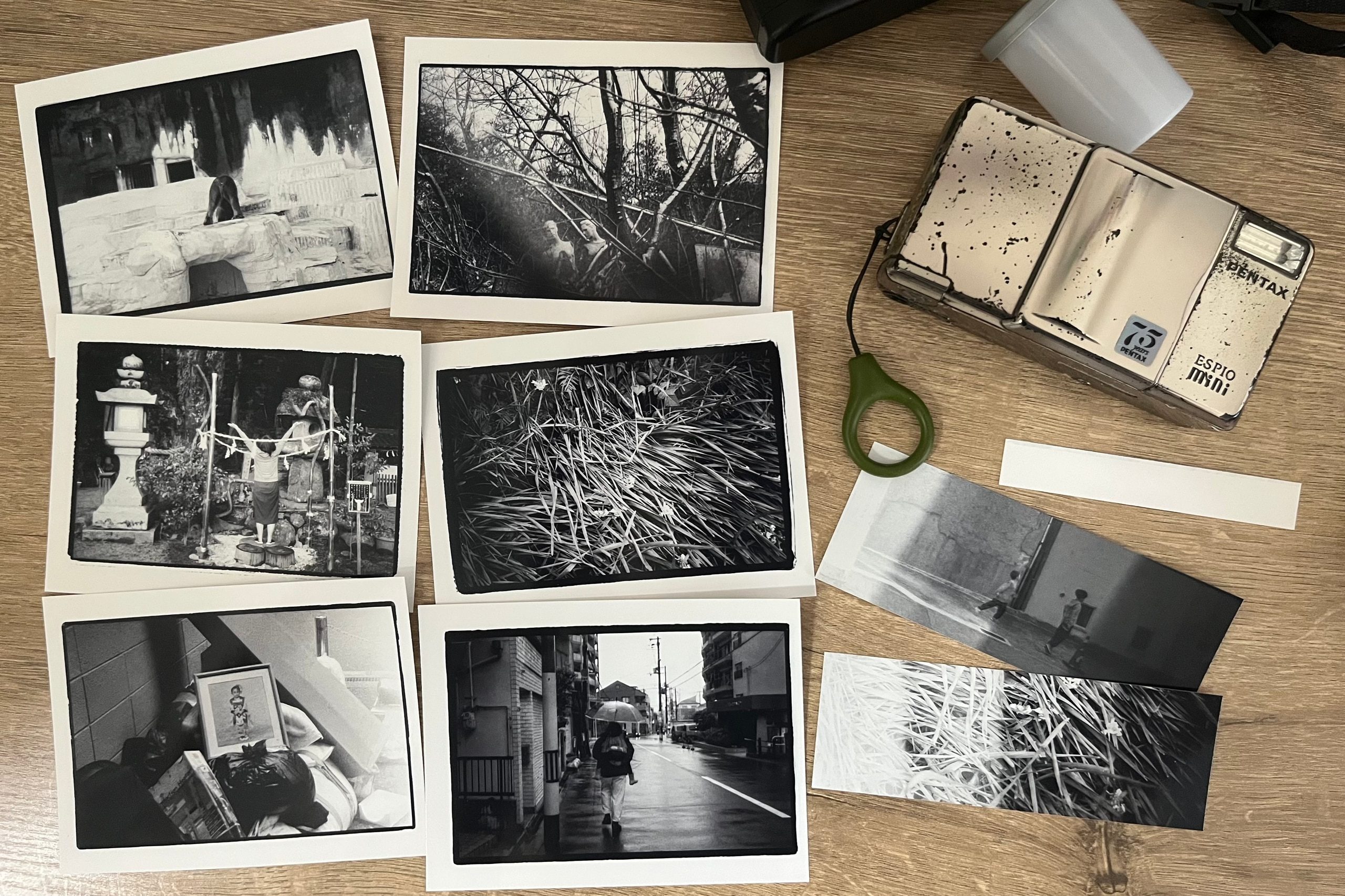 Black-and-white prints laid out on a wooden table beside a vintage Pentax Espio Mini film camera and two contact-print strips.