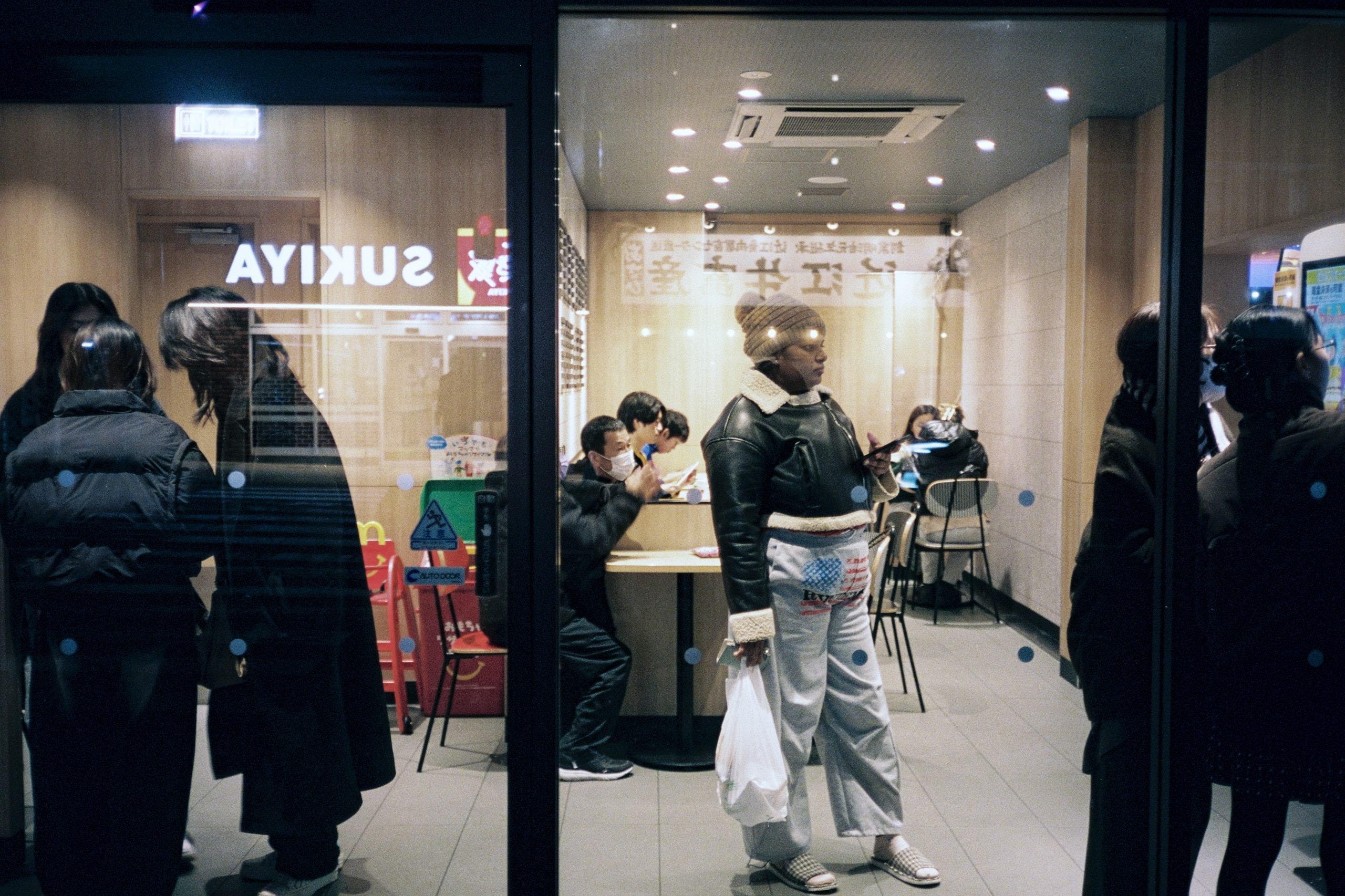 People wait outside a restaurant window at night, reflections layered across the glass.