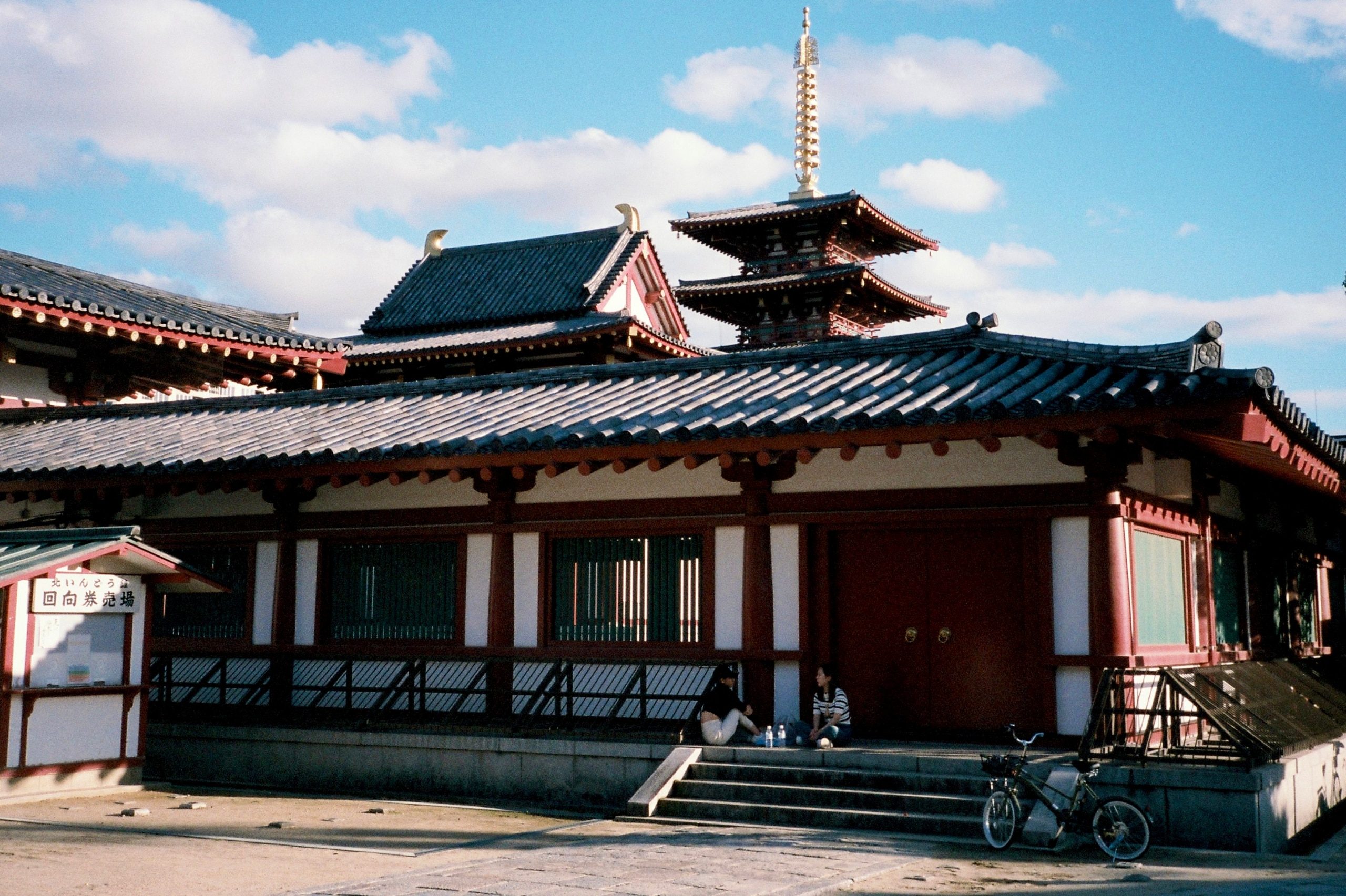 A temple's tiled roof and pagoda spire against a blue sky with clouds; two people sit at its base.