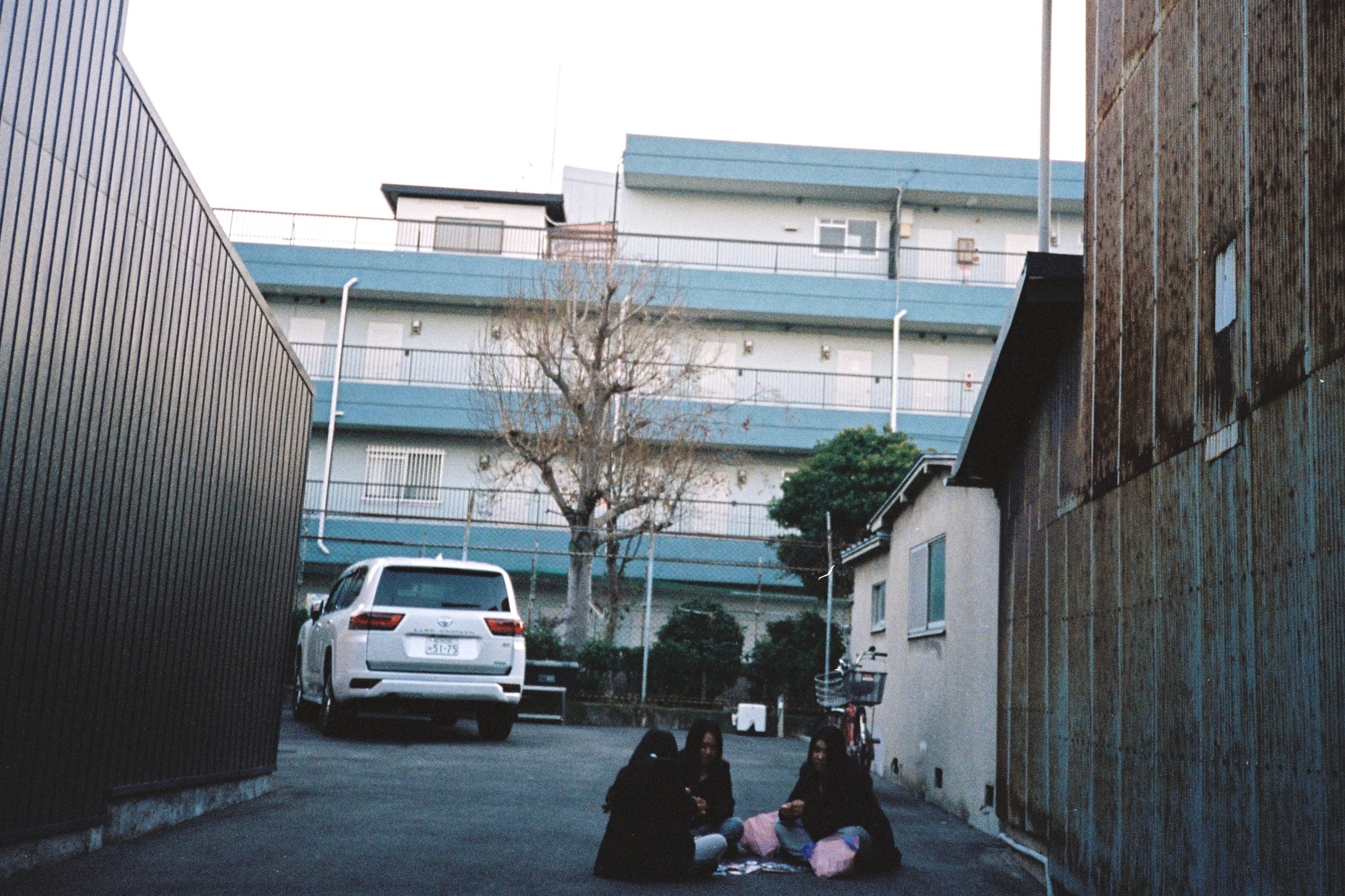 Three people sit together in a parking lot alley, apartment blocks behind them.