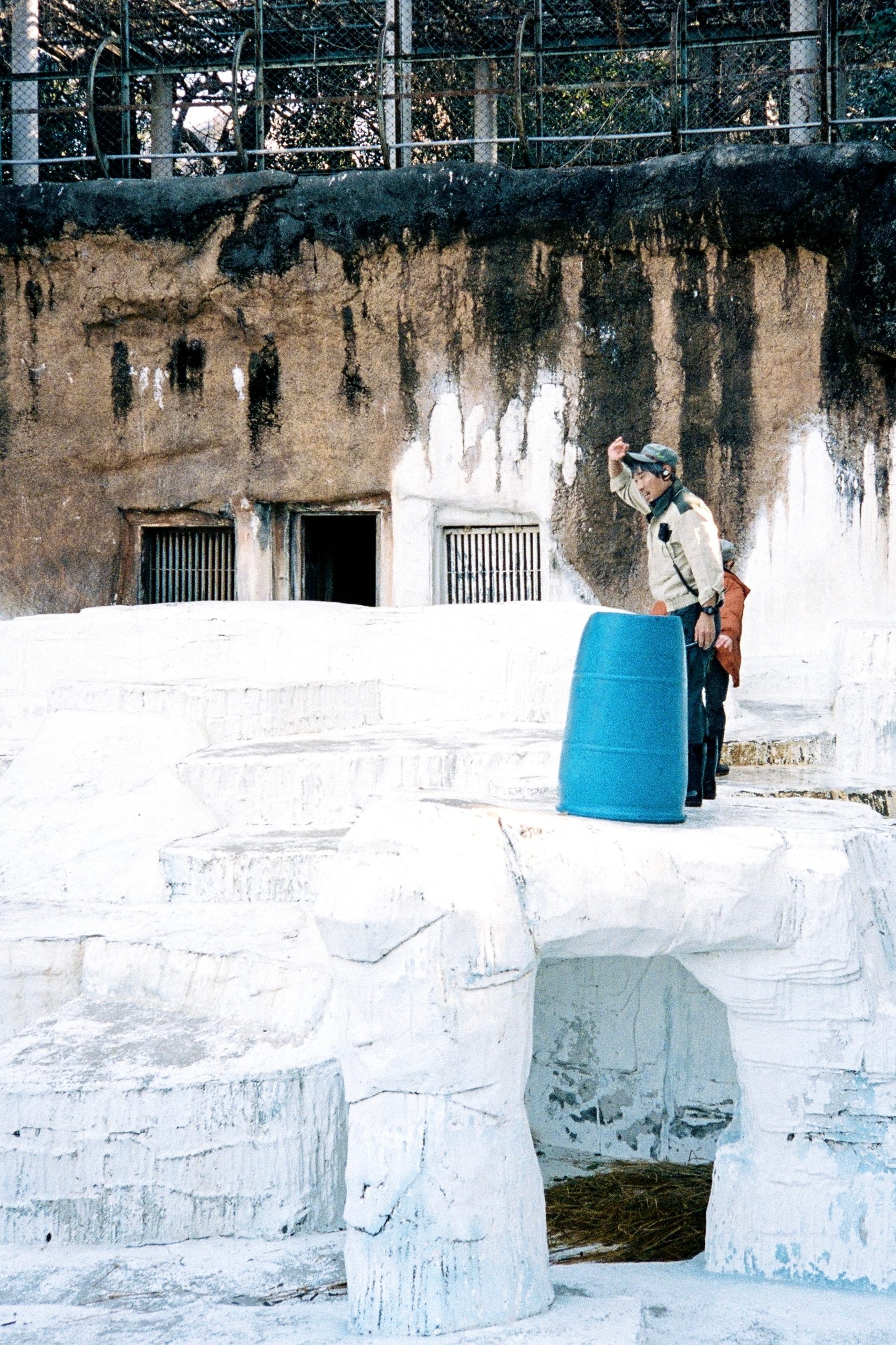 A zookeeper stands alone in an empty polar bear enclosure beside a blue barrel.