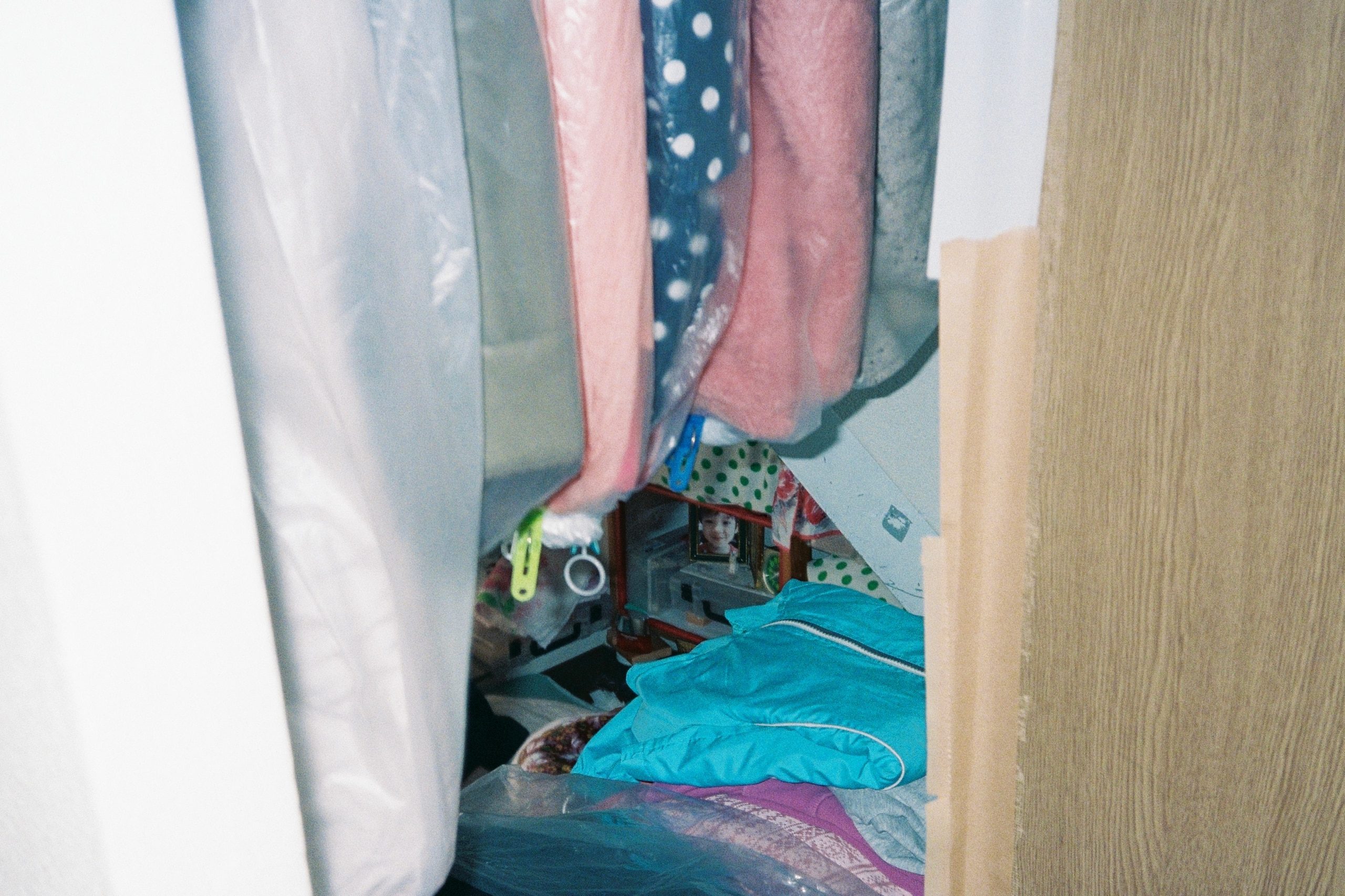 A cluttered closet seen between hanging clothes in plastic covers, a small framed photo visible behind.