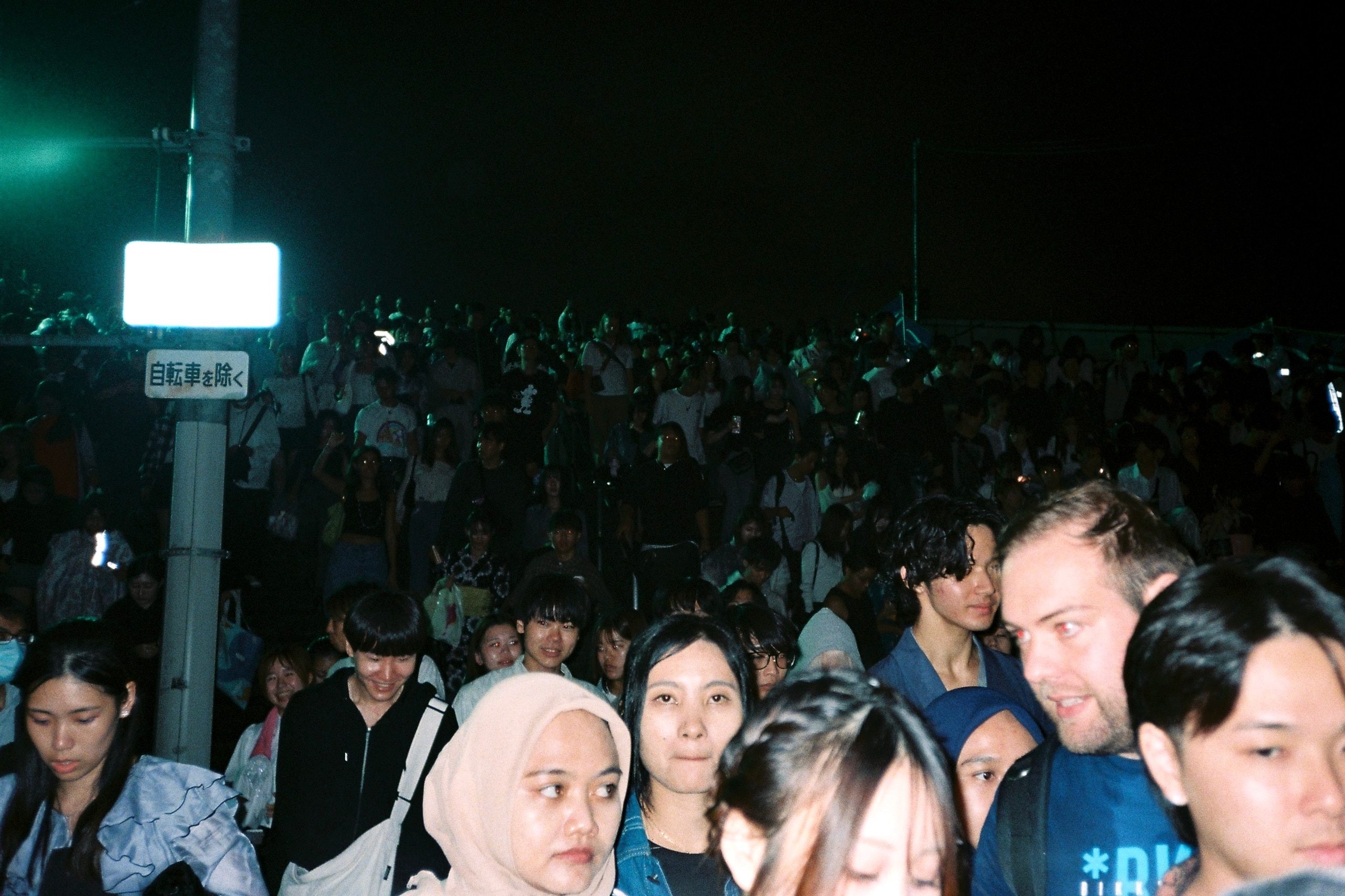 A dense nighttime crowd fills a street, faces lit by nearby signs and phone screens.