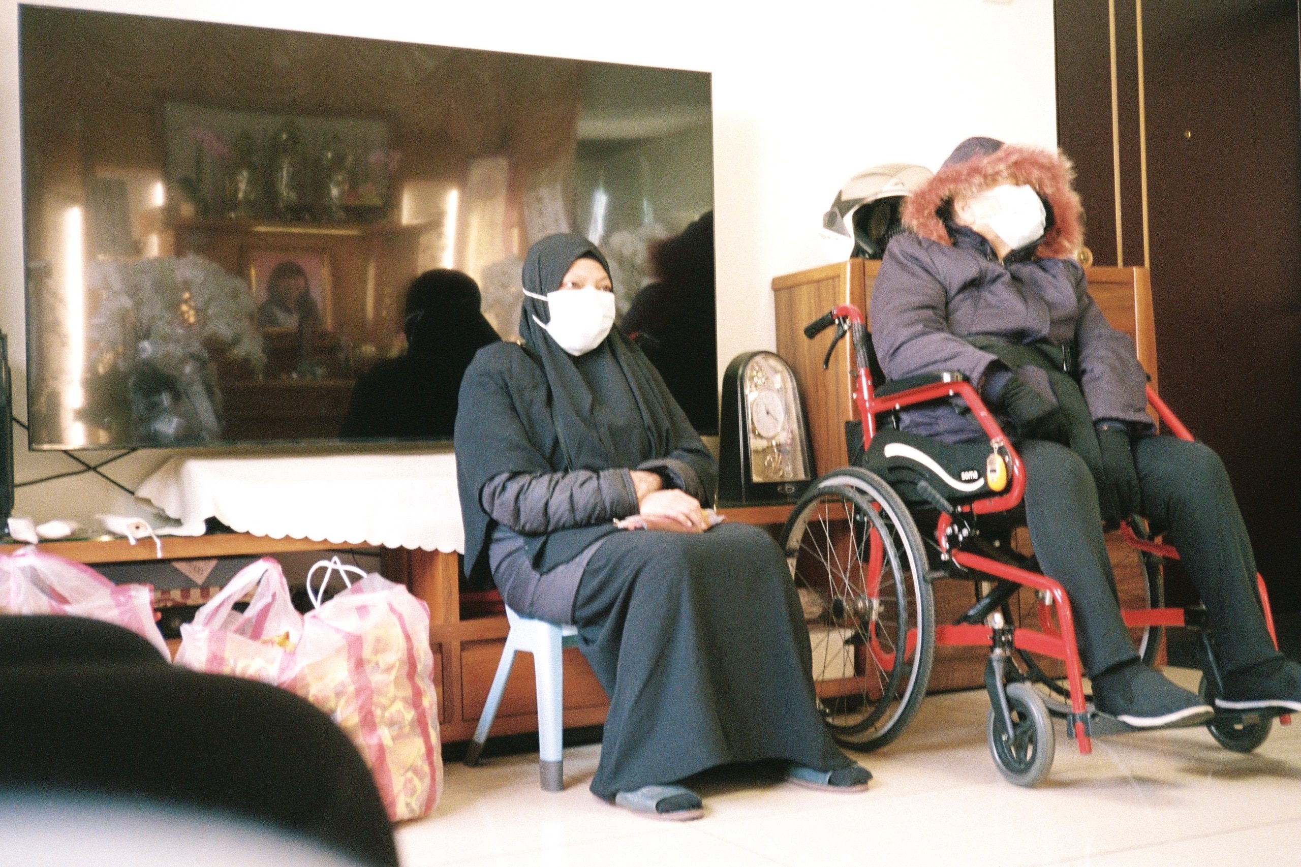 Two masked people wait in a living room, one seated, one in a wheelchair, shopping bags on the floor.