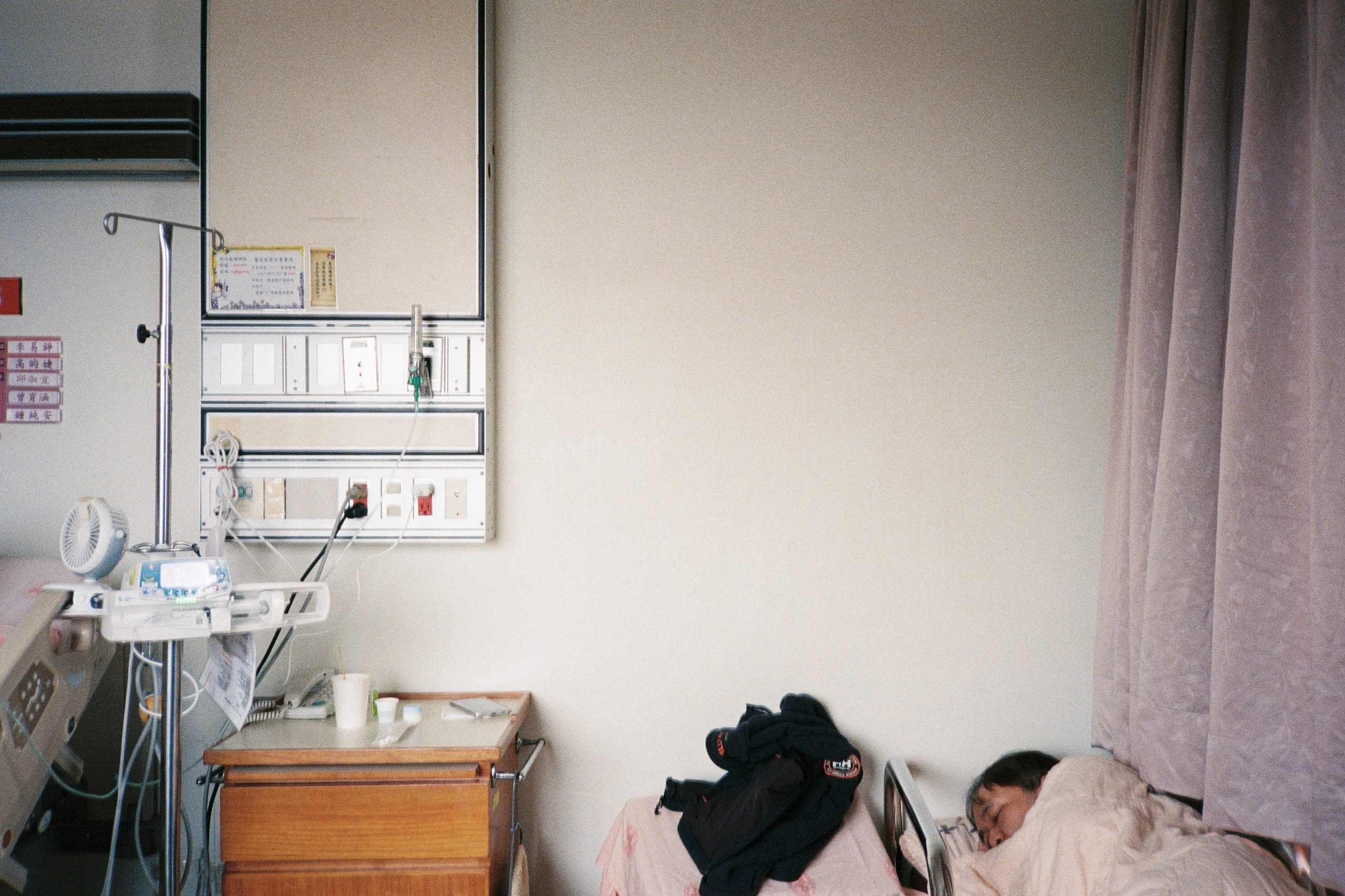 A hospital room with a patient sleeping in bed, an IV pole beside a large blank wall.