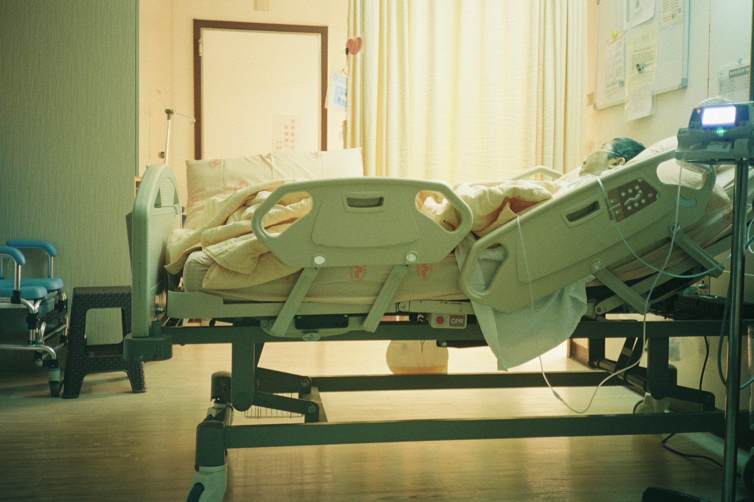 A hospital bed lit by a sunlit curtain, a patient resting as a monitor glows nearby.