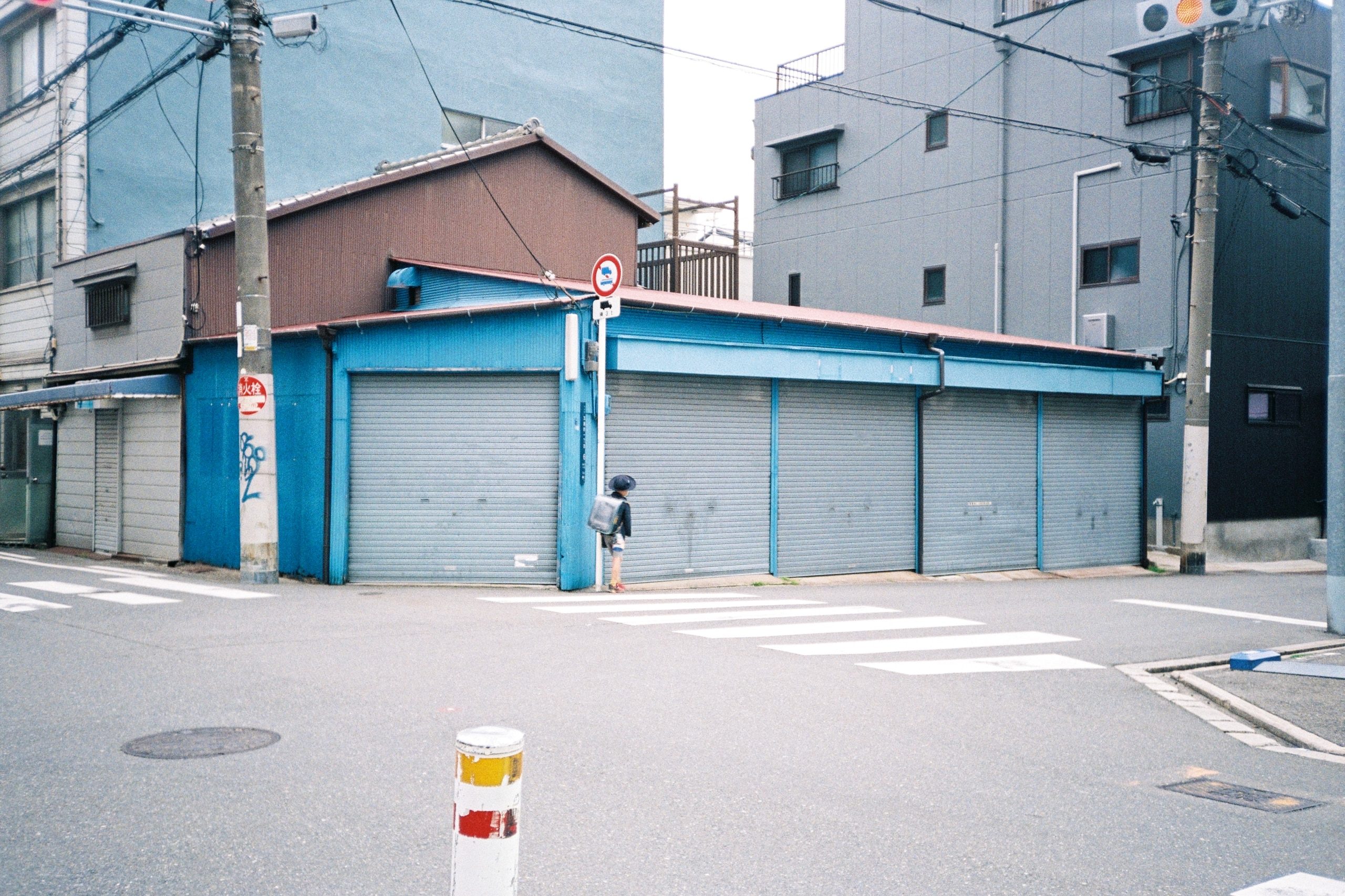 A child with a school backpack walks alone past a row of closed blue shutters at a street corner.